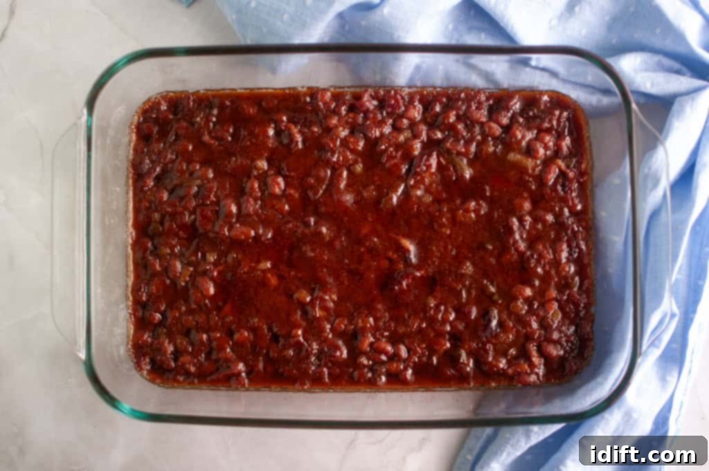 A glass baking dish filled with baked beans in a reddish-brown sauce, placed on a light countertop next to a blue cloth, as it bakes to bubbly perfection.
