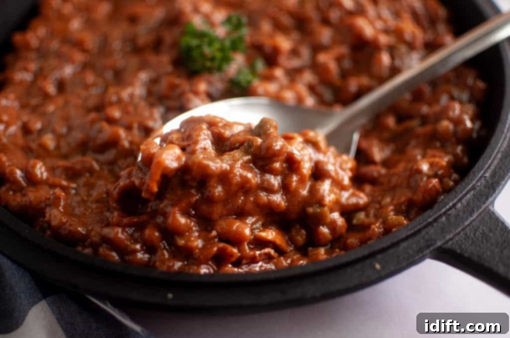 Homestyle Bean Bake 13 A close-up shot of a cast-iron skillet brimming with rich, bubbling baked beans, adorned with a delicate sprig of fresh parsley. A metal spoon is poised, lifting a generous portion, highlighting the thick, saucy texture and inviting flavor of this comforting dish.