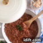 A white Dutch oven with baked beans topped with parsley sits on a marble surface next to a jar of brown sugar, a wooden spoon, and a blue cloth. This rich, smoky baked beans casserole is ready to be served.