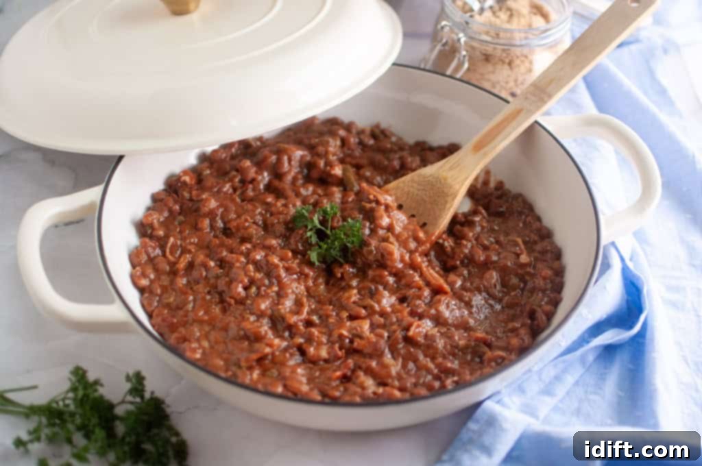 A pot of cooked pinto beans topped with parsley sits on a counter with a wooden spoon inside and a jar of brown sugar in the background, showcasing the fresh ingredients for homemade baked beans.