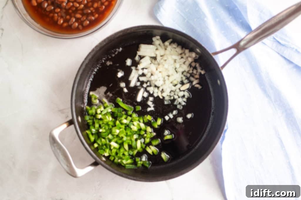 A saucepan on a stovetop contains chopped onions and green peppers in oil, ready to be cooked. A bowl of beans and a blue cloth are nearby, illustrating the next step in the recipe.