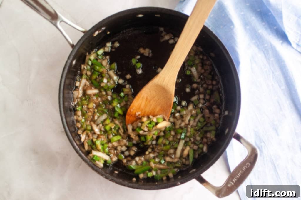 Chopped onions and green peppers are being sautéed in a black saucepan with a wooden spoon on a light countertop, cooking down to tender perfection for the baked beans.