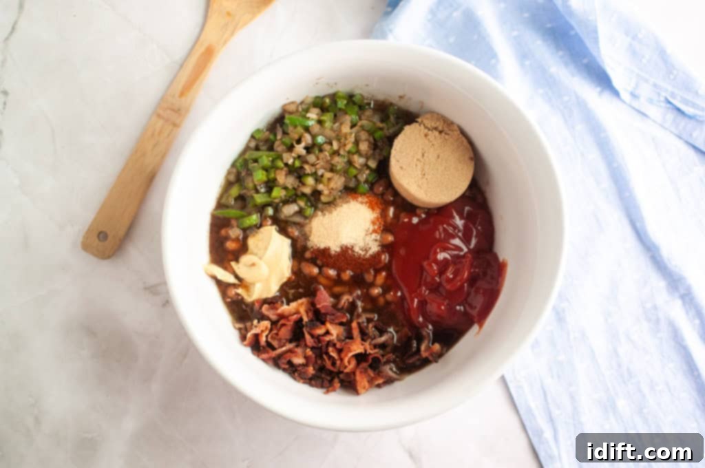 A white bowl with chopped green peppers, brown sugar, ketchup, bacon, beans, seasonings, and butter on a marble surface next to a wooden spoon and a blue cloth, indicating all components are ready for mixing.