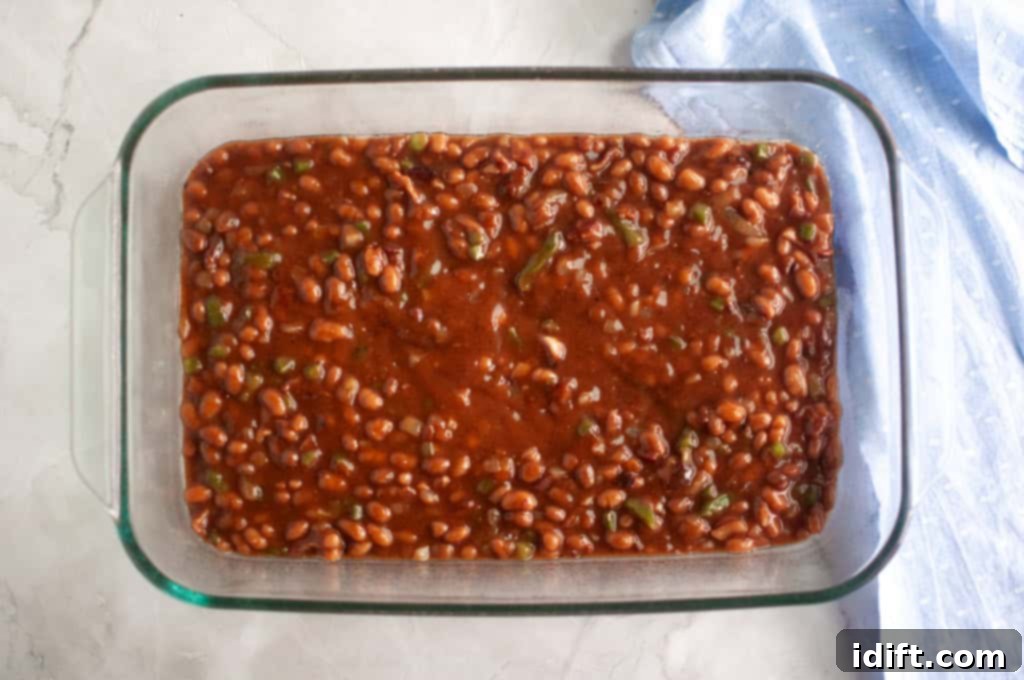 A glass baking dish filled with baked beans in sauce sits on a light countertop next to a folded blue cloth, prepped and ready for the oven.