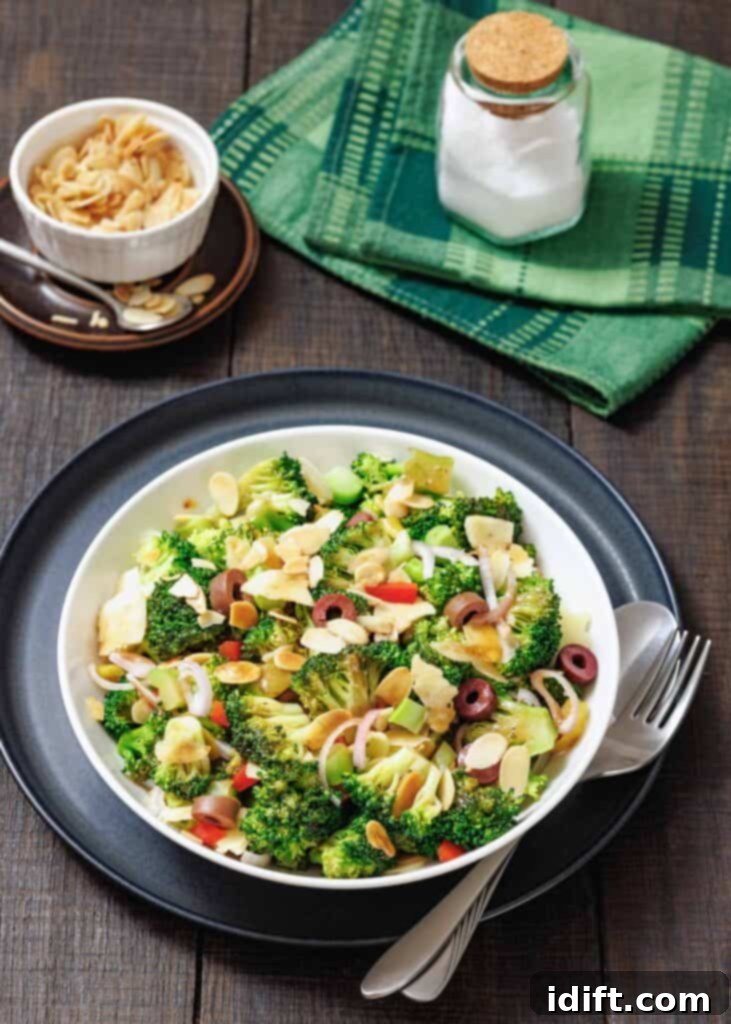 A bowl of Italian Broccoli Salad with sliced almonds, red onion, olives, and cheese, served with a fork and spoon on a dark plate; salt and napkin in the background.