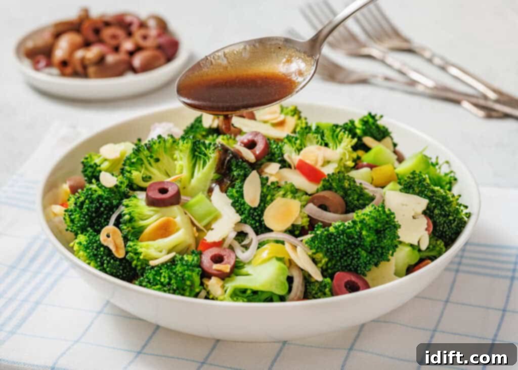 A bowl of broccoli salad with olives, sliced almonds, and vegetables is being drizzled with dressing from a spoon. A plate of olives and forks are in the background.