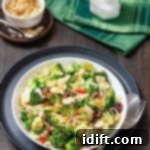 A bowl of broccoli salad with sliced almonds, red onion, olives, and cheese, served with a fork and spoon on a dark plate; salt and napkin in the background.