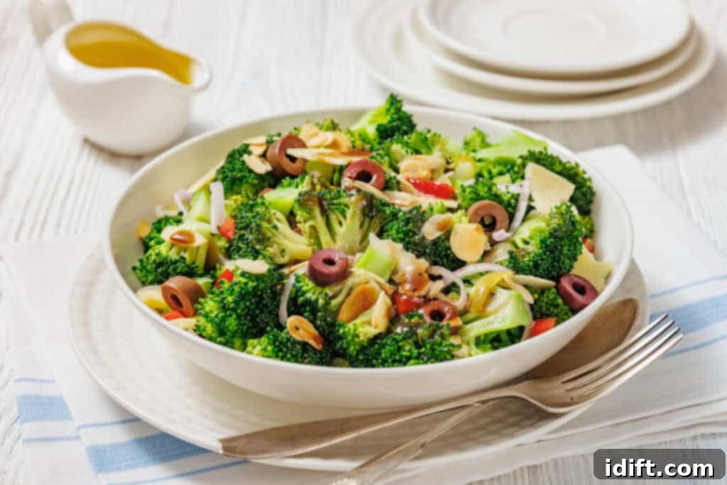 A bowl of broccoli salad with sliced olives, almonds, onions, and red peppers, served on a white plate with a fork and napkin beside it. A small pitcher of dressing is in the background.
