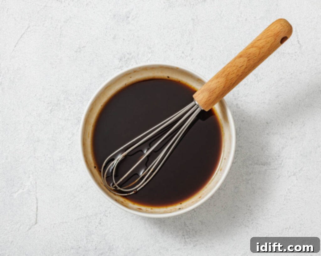 A metal whisk rests in a white bowl filled with a dark liquid, possibly a sauce or marinade, on a light textured surface.