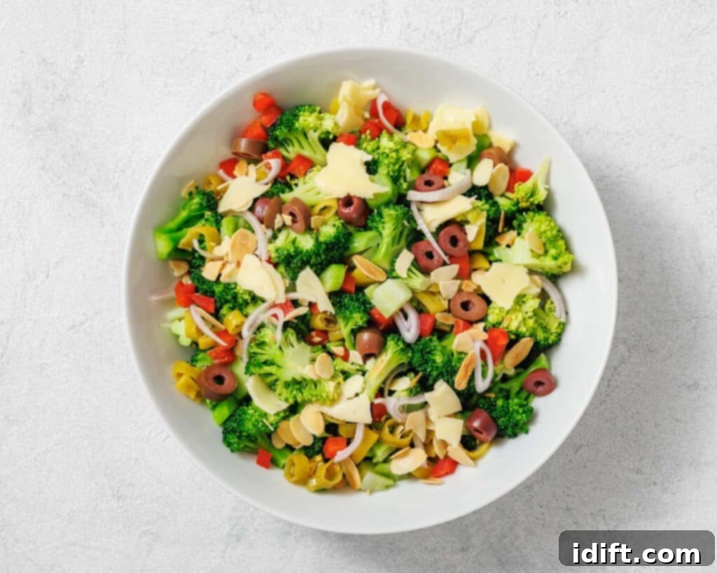 A bowl of broccoli salad with sliced olives, red bell peppers, onions, shaved cheese, and slivered almonds on a light surface.