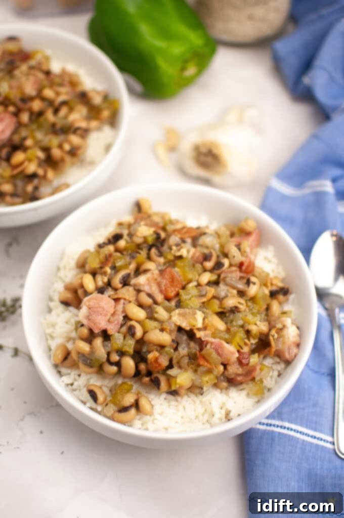 A bowl of rice topped with black-eyed peas, diced ham, and vegetables sits on a marble surface next to a spoon, a green bell pepper, garlic, and a blue napkin.