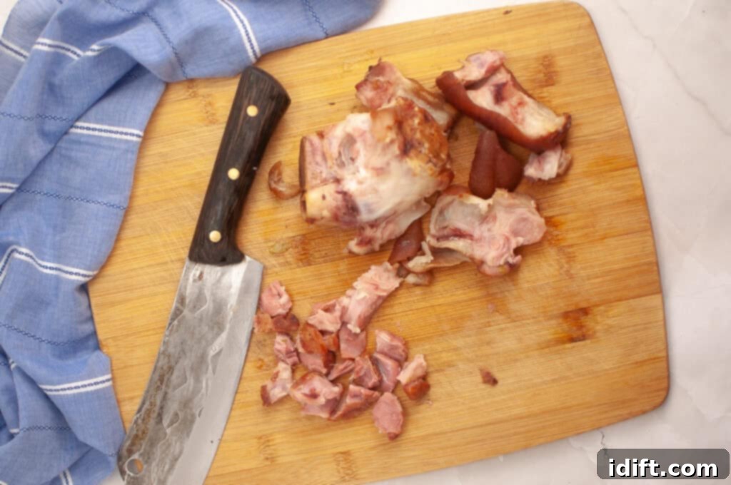A wooden cutting board with a large knife, a blue kitchen towel, and chopped pieces of smoked ham hock.