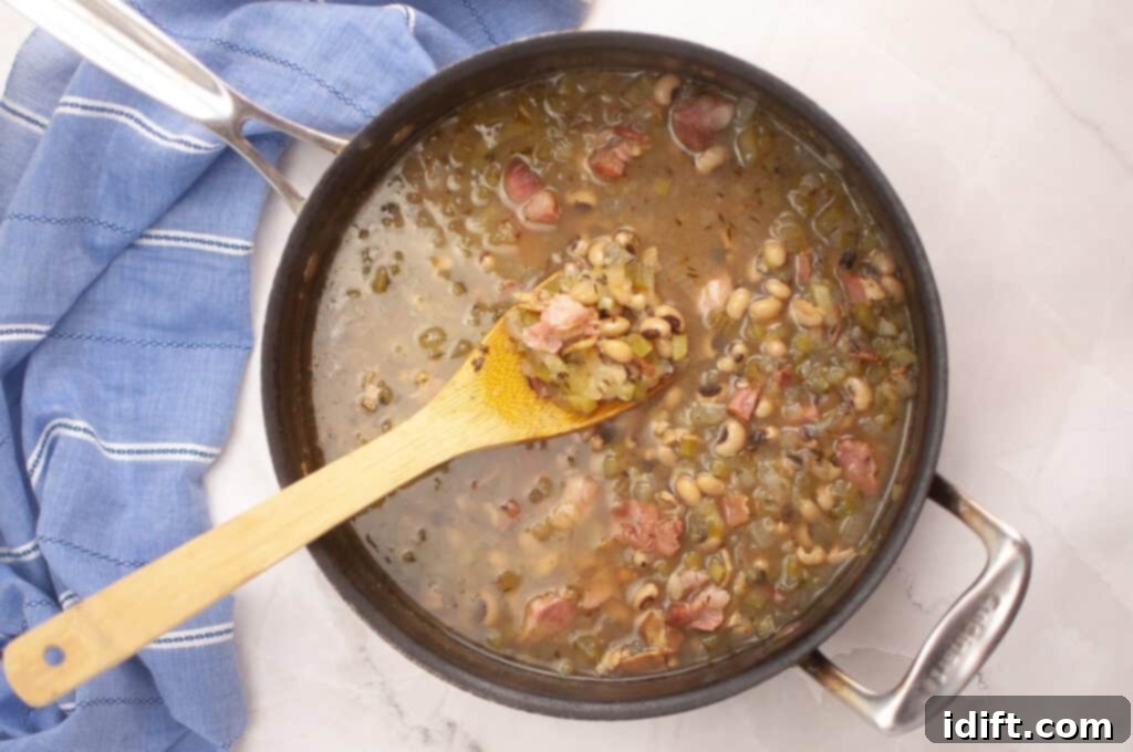 A pot of black-eyed peas with ham and vegetables simmers, with a wooden spoon resting inside; a blue kitchen towel is beside the pot on a white marble surface.