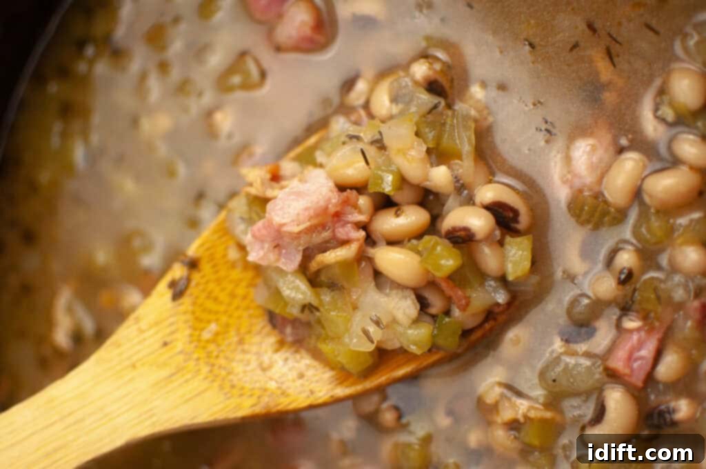 A wooden spoon holds black-eyed peas, diced vegetables, and pieces of ham in a broth.