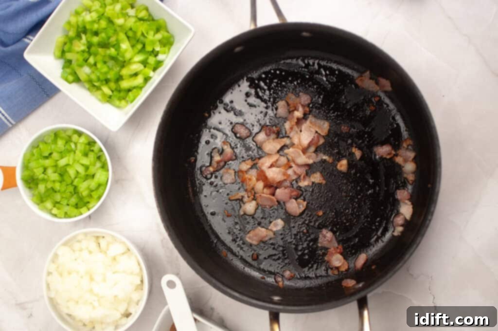 Chopped bacon cooking in a black pan, with bowls of diced celery, green bell pepper, and onion arranged nearby on a white countertop.