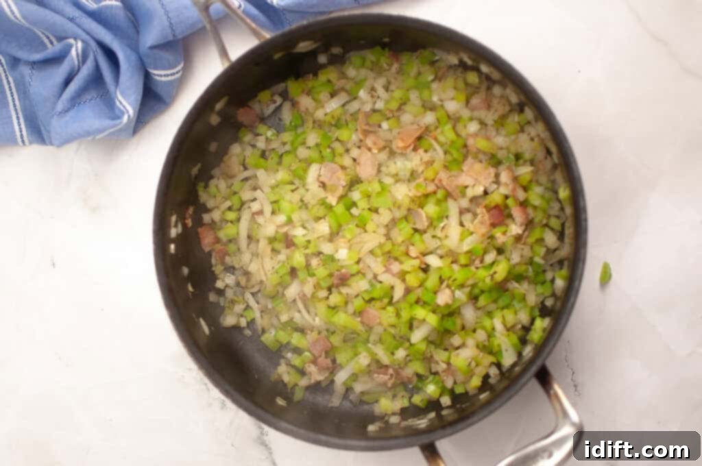A pot containing chopped celery, onions, and bacon pieces being sautéed on a stovetop, with a blue kitchen towel nearby.