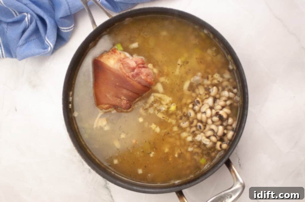 A pot of soup with a large piece of smoked meat and black-eyed peas simmering in broth on a white surface, next to a blue cloth.