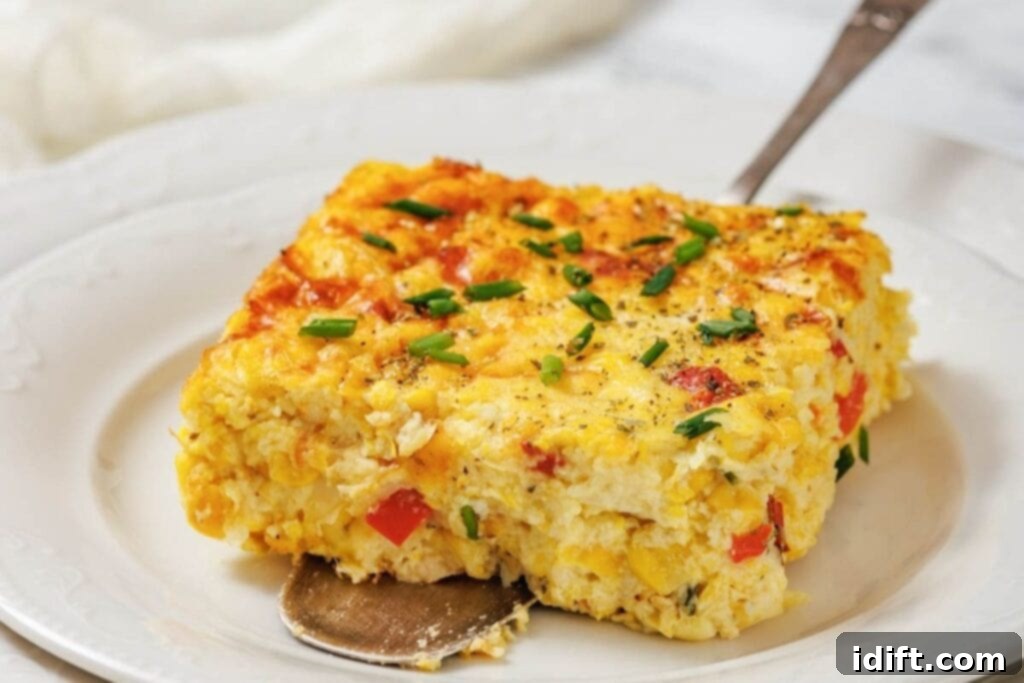 Creamy Golden Corn Bake 3 A square slice of corn pudding casserole with eggs, red bell pepper, and chives sits on a white plate with a serving spoon underneath.