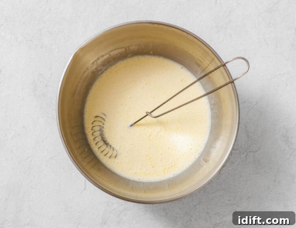 Creamy Golden Corn Bake 6 A whisk rests in a metal bowl containing a pale, mixed liquid batter on a light gray surface.
