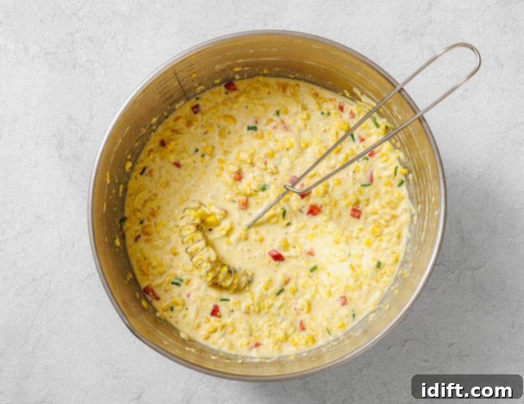 Creamy Golden Corn Bake 8 A mixing bowl filled with a creamy batter containing corn, red bell pepper, green onions, and a whisk resting inside.