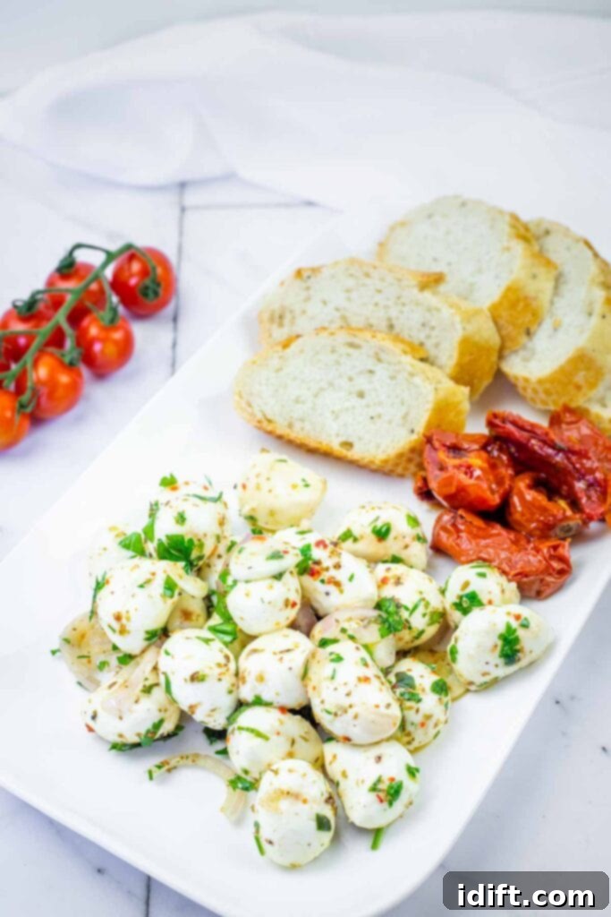 A rectangular white plate with sliced bread, marinated mozzarella balls with herbs, roasted tomatoes, and a vine of cherry tomatoes on the side.