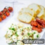 A rectangular white plate with sliced bread, marinated mozzarella balls with herbs, roasted tomatoes, and a vine of cherry tomatoes on the side.