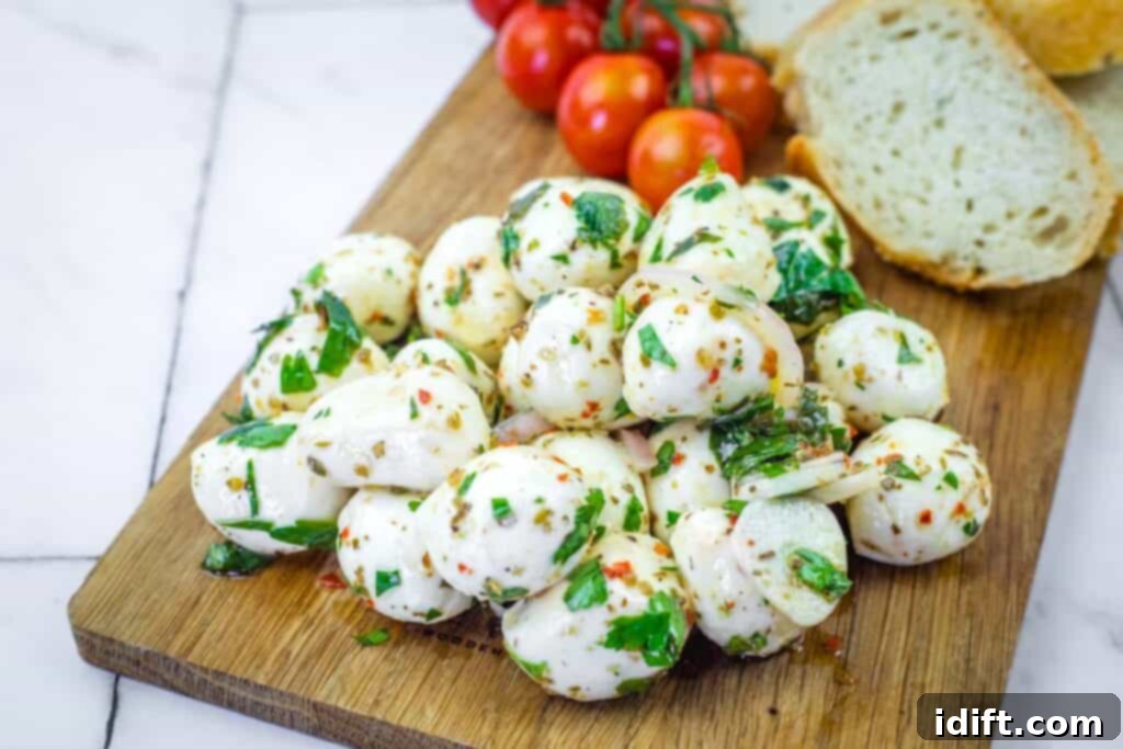 A pile of marinated mozzarella balls garnished with herbs on a wooden board, with cherry tomatoes and slices of bread in the background.