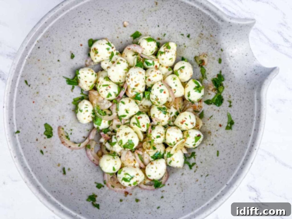 A speckled bowl filled with mozzarella balls, sliced red onions, chopped herbs, and seasoning on a white marble surface.