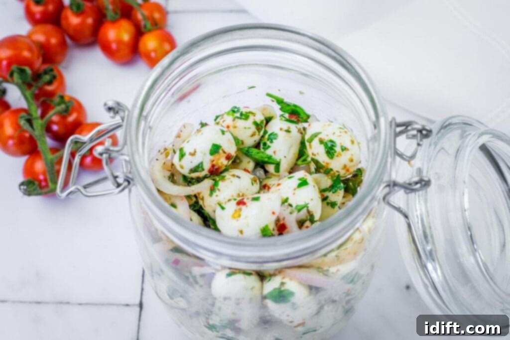 A glass jar filled with marinated mozzarella balls, onion slices, and herbs sits on a marble surface next to a vine of cherry tomatoes.