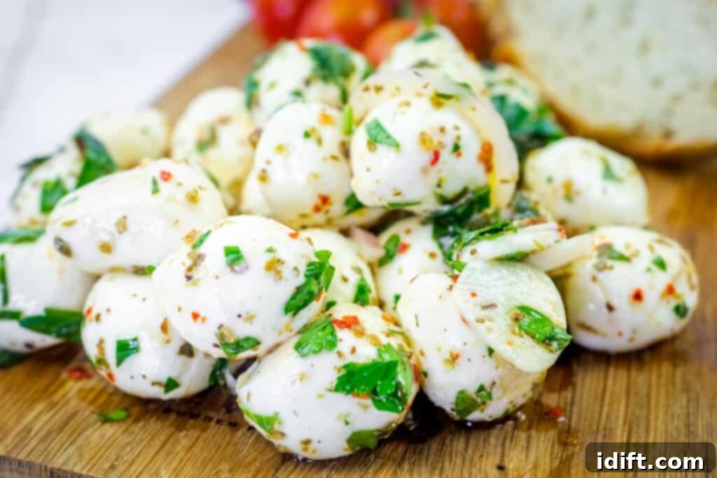 A pile of marinated mozzarella balls with herbs and spices on a wooden board, with tomatoes and bread in the background.