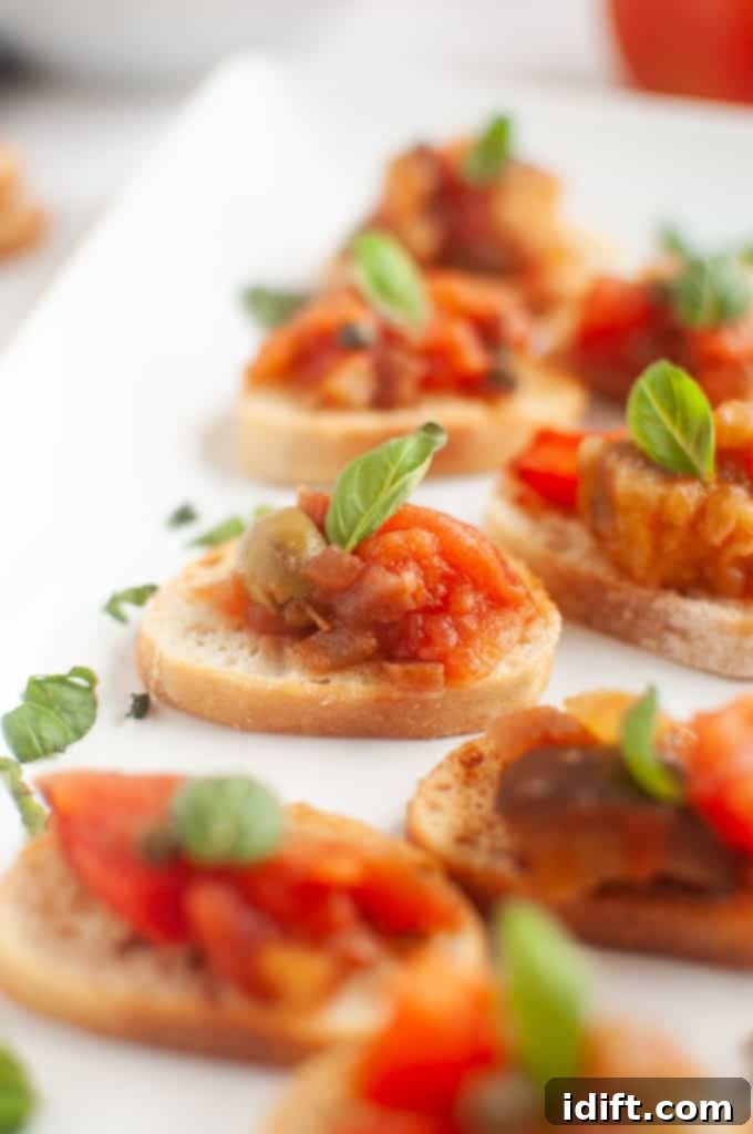 A white rectangular plate with several slices of toasted bread topped with Eggplant Caponata and fresh basil leaves.
