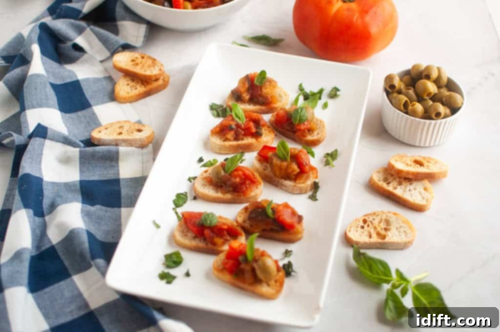 A white rectangular plate with several pieces of toasted bread topped with Eggplant Caponata and fresh basil, surrounded by ingredients.