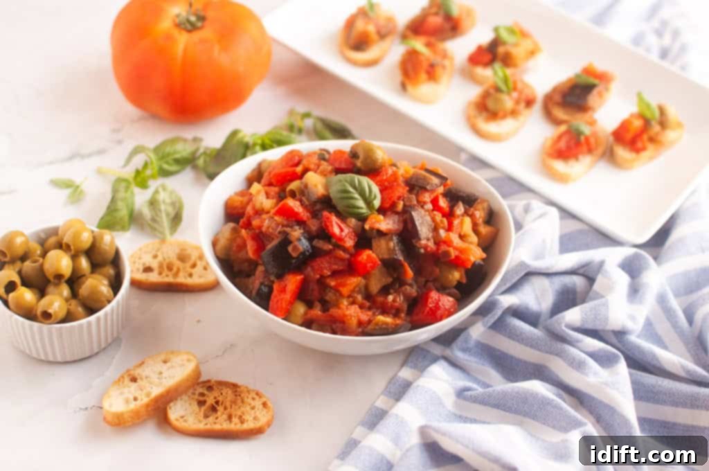 A bowl of Eggplant Caponata, garnished with basil, is surrounded by toasted bread, olives, a tomato, and bruschetta.