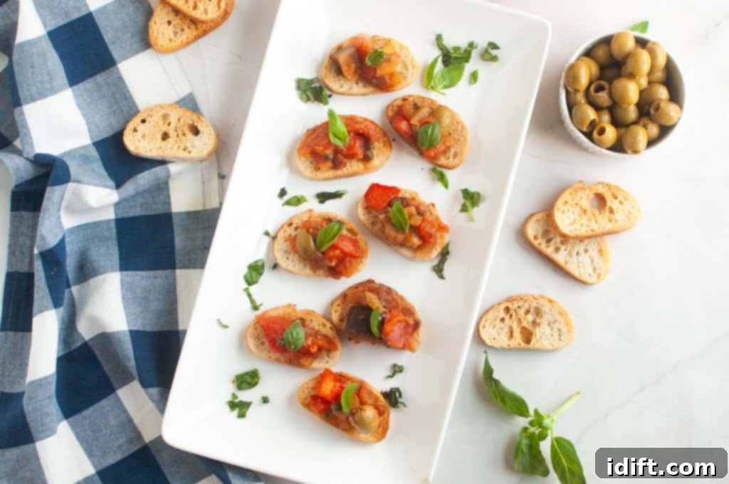 A white rectangular plate with several pieces of toasted bread topped with Eggplant Caponata and fresh basil, with olives and extra bread.