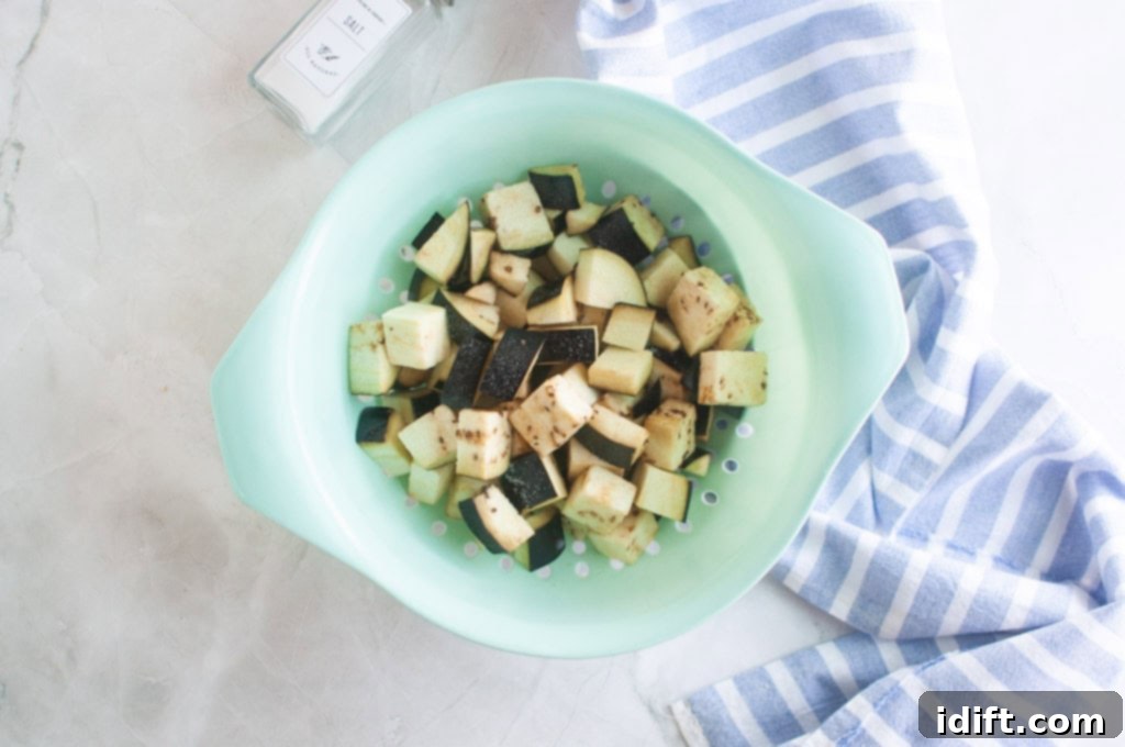 Cubed eggplant pieces in a light green colander, next to a salt container and a blue and white striped dish towel on a light surface.