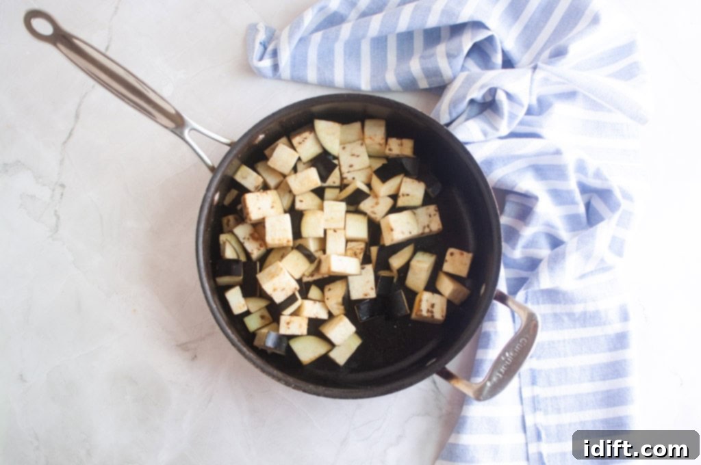 A black saucepan containing cubed eggplant sits on a light countertop next to a blue and white striped cloth.