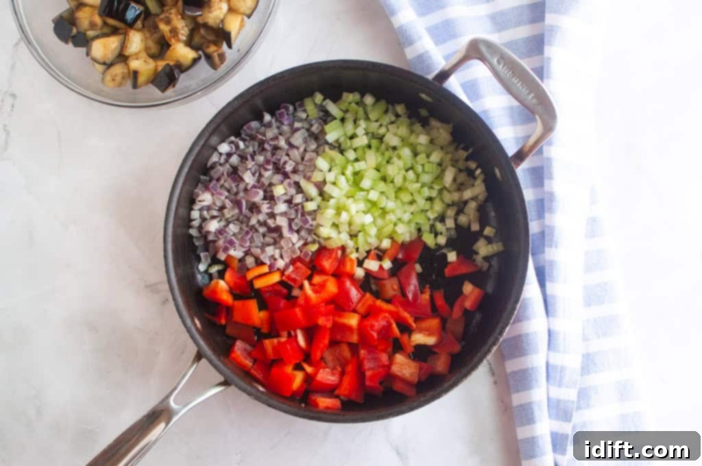 A skillet with diced red bell pepper, celery, and red onion, with a bowl of cooked eggplant and a striped towel beside it on a marble surface.