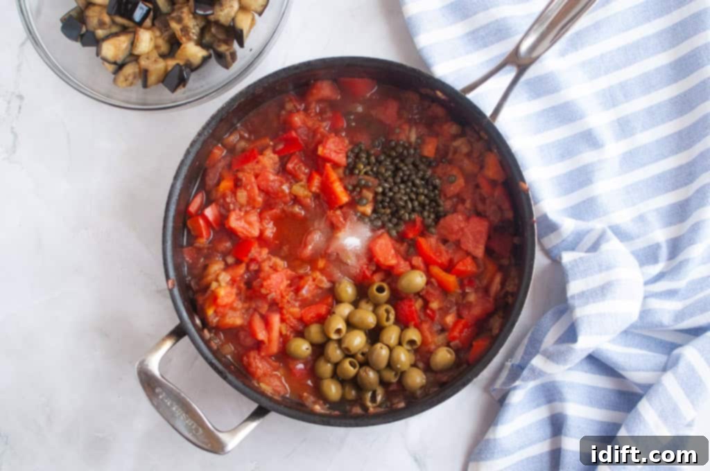 A pan filled with chopped tomatoes, green olives, capers, and vegetables sits on a counter next to a bowl of diced eggplant and a striped kitchen towel.