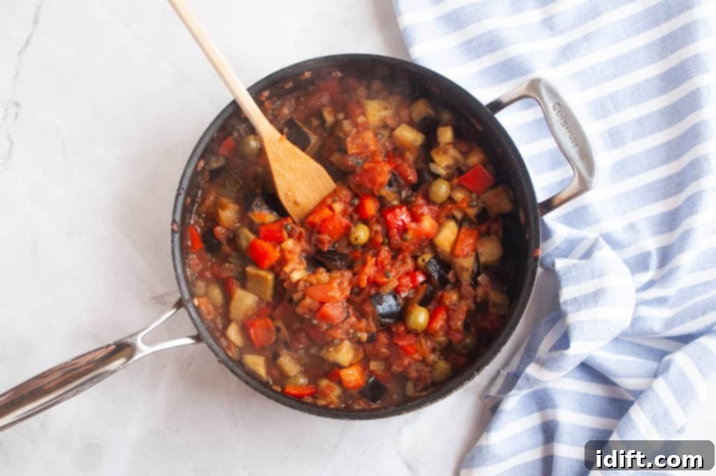 A pan of vegetable stew with tomatoes, eggplant, and olives sits on a marble surface next to a blue-and-white striped towel; a wooden spoon rests in the pan.