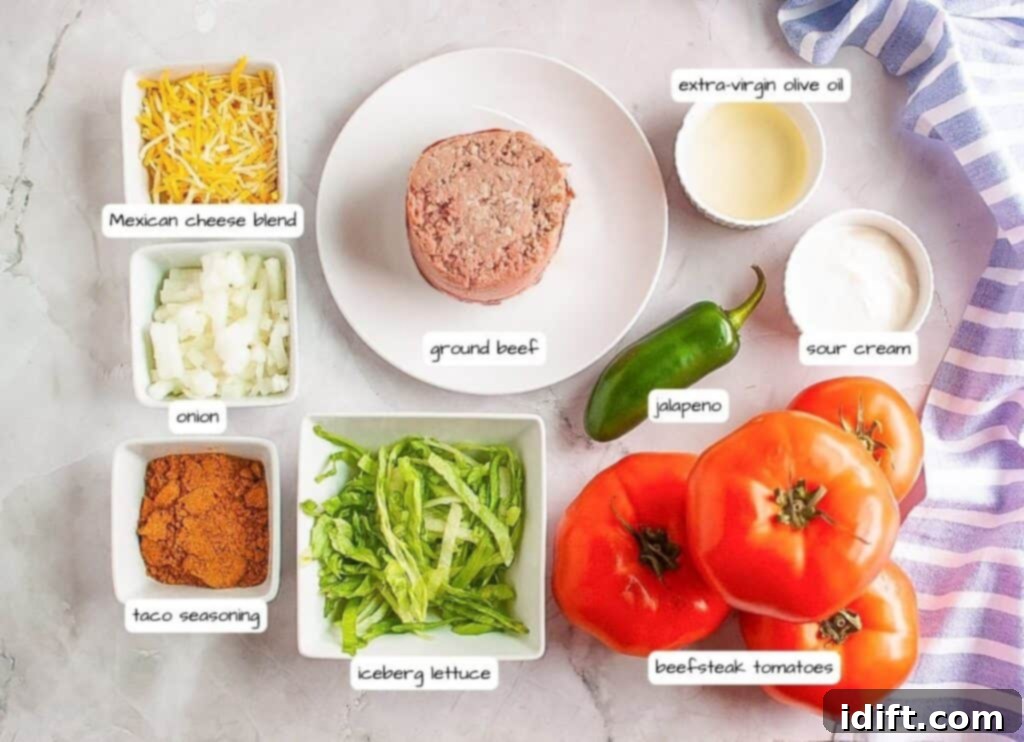 An overhead shot showcasing all the labeled ingredients meticulously laid out for the taco stuffed tomatoes recipe, including ground beef, fresh tomatoes, cheese, onions, lettuce, taco seasoning, jalapeños, olive oil, and sour cream.
