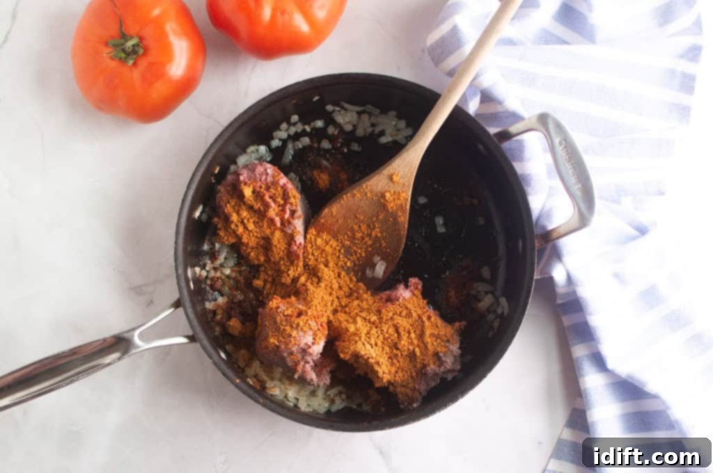 An overhead shot of ground beef, diced onions, and vibrant taco spices being skillfully stirred together in a black skillet with a wooden spoon.