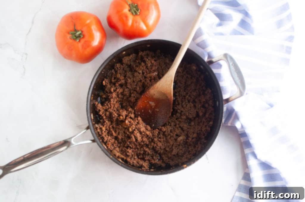 An overhead shot of well-seasoned ground meat being stirred in a black skillet with a sturdy wooden spoon, ensuring even browning and rich flavor.