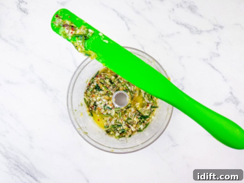 Aromatic Slow-Roasted Lamb Leg 7 Overhead shot of finely chopped herbs and spices for chimichurri in a food processor, with a green spatula nearby, showing the consistency of the paste.