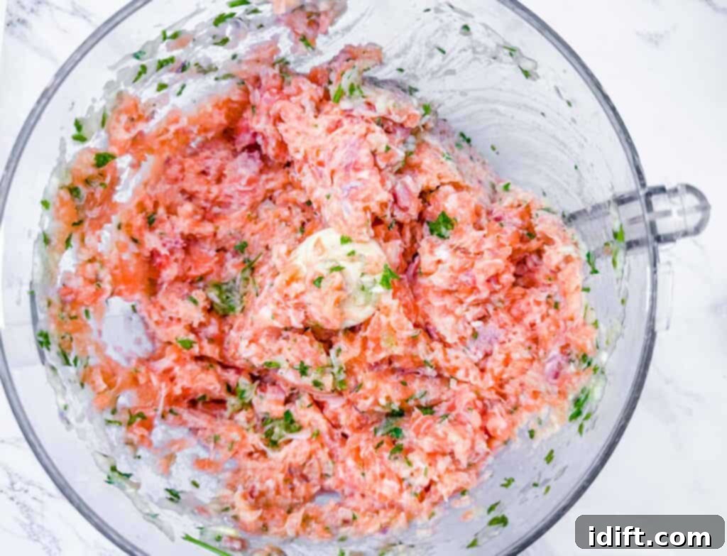 Overhead shot of raw salmon chunks being gently processed with fresh herbs and other aromatic ingredients in a food processor, just until finely chopped, not puréed.
