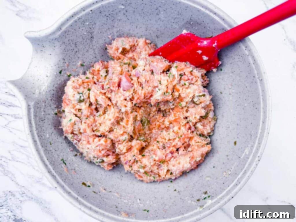 Overhead shot of the vibrant raw salmon mixture being gently stirred with a red spatula in a sleek gray mixing bowl, showcasing the added Panko, mayonnaise, shallots, parsley, capers, lemon juice, Dijon, salt, and pepper.