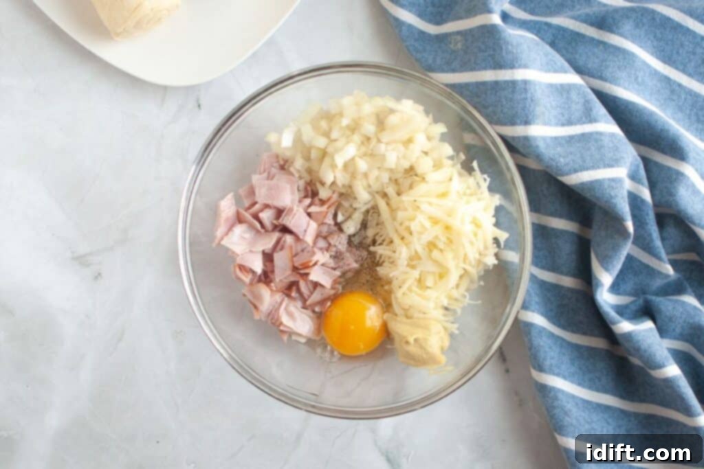 Fluffy Ham and Cheese Bites 6 Overhead shot of a glass bowl containing diced ham, shredded Swiss cheese, diced onion, a raw egg, Dijon mustard, and a sprinkle of pepper, ready to be mixed.