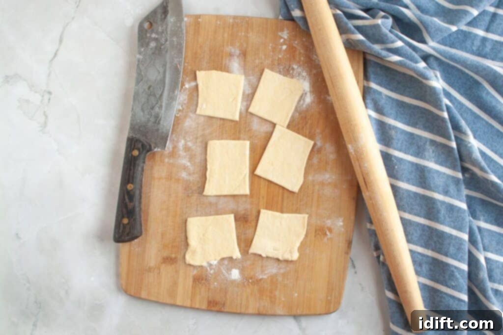 Fluffy Ham and Cheese Bites 7 Overhead shot of unrolled crescent dough being cut into small, uniform square pieces on a wooden board using a pizza cutter, with a rolling pin nearby.