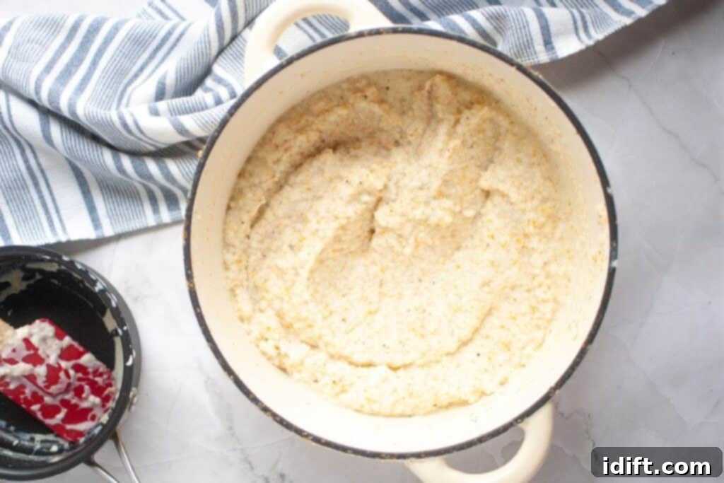 A pot of cooked grits sits on a white countertop with a blue and white striped towel nearby and a used spatula resting in a small black bowl.
