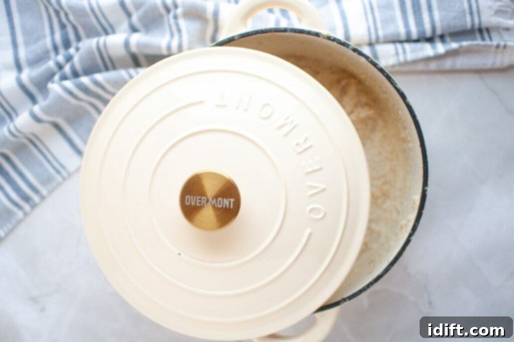 A cream-colored Overmont Dutch oven with a partially open lid reveals cooked rice inside, placed on a white surface with a blue and white striped cloth nearby.