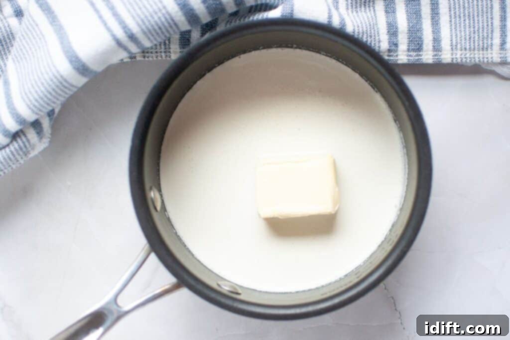 A saucepan containing milk and a block of butter, next to a striped kitchen towel on a white surface.